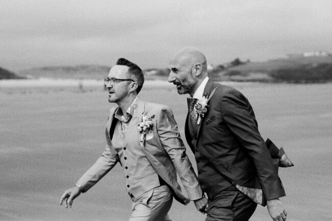 Two grooms walking hand in hand on a beach during their wedding in Cork, Ireland